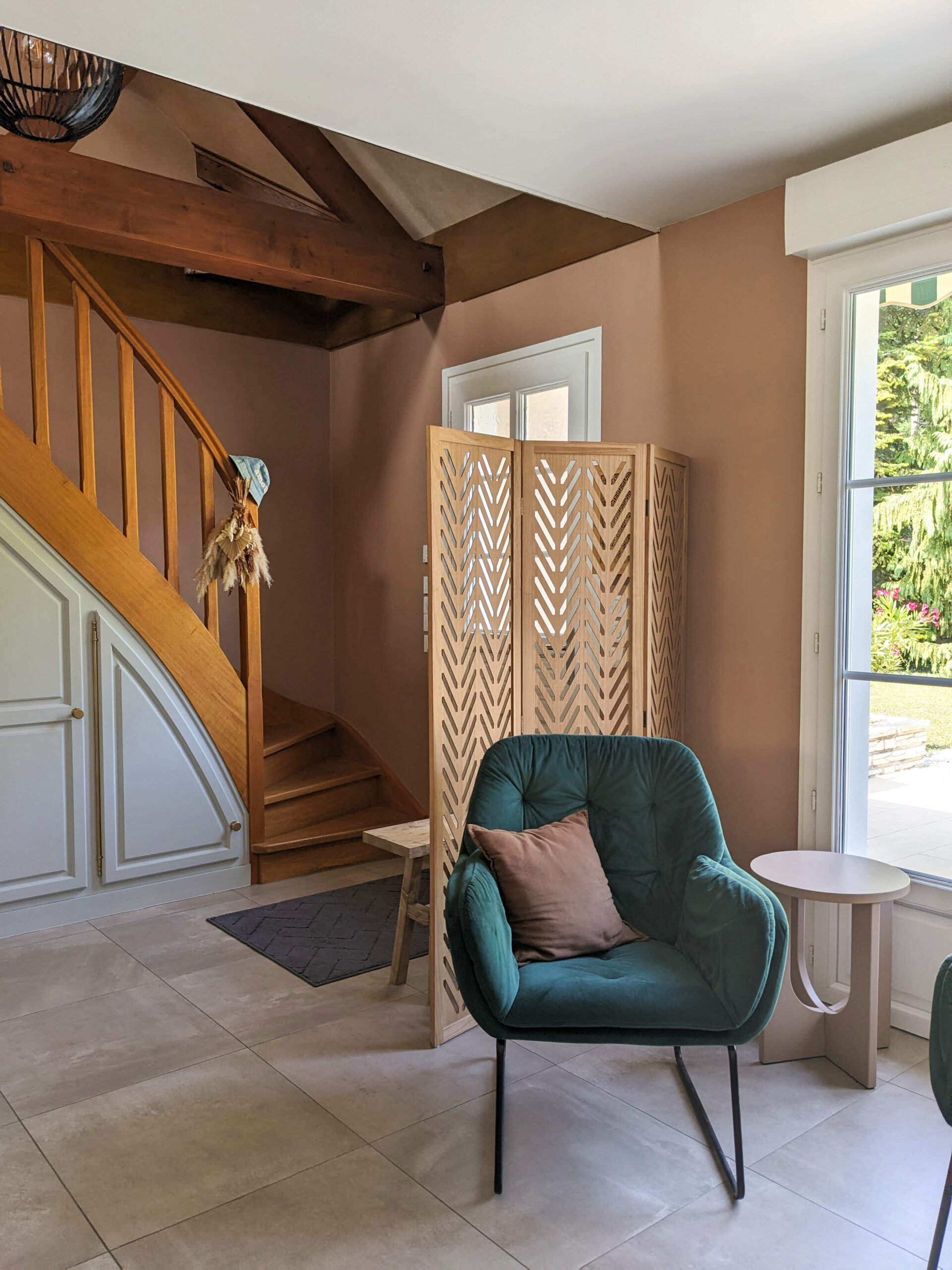 Entrée de maison avec poutres en bois apparentes, suspension boule en bois, paravent et banc en bois, mur terracotta et fauteuil en velours vert sapin Dans une maison familiale en campagne essonnienne à Chalo-Saint-Mars, vue sur l’entrée avec poutres en bois apparentes, suspension boule en bois, paravent et banc en bois, mur terracotta et fauteuil en velours vert sapin