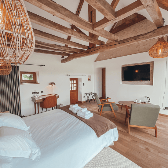 Domaine de Prestal rénovation ancien four à pain en chambre d'hotes, coin salon et nuit avec bureau Réhabilitation d'un ancien four à pain en chambre d'hôtes chaleureuse avec poutres apparentes, mur en pierre et lumière naturelle, fauteuil en bois massif et tissu vert kaki dans le coin salon, au Merlerault dans le perche normand