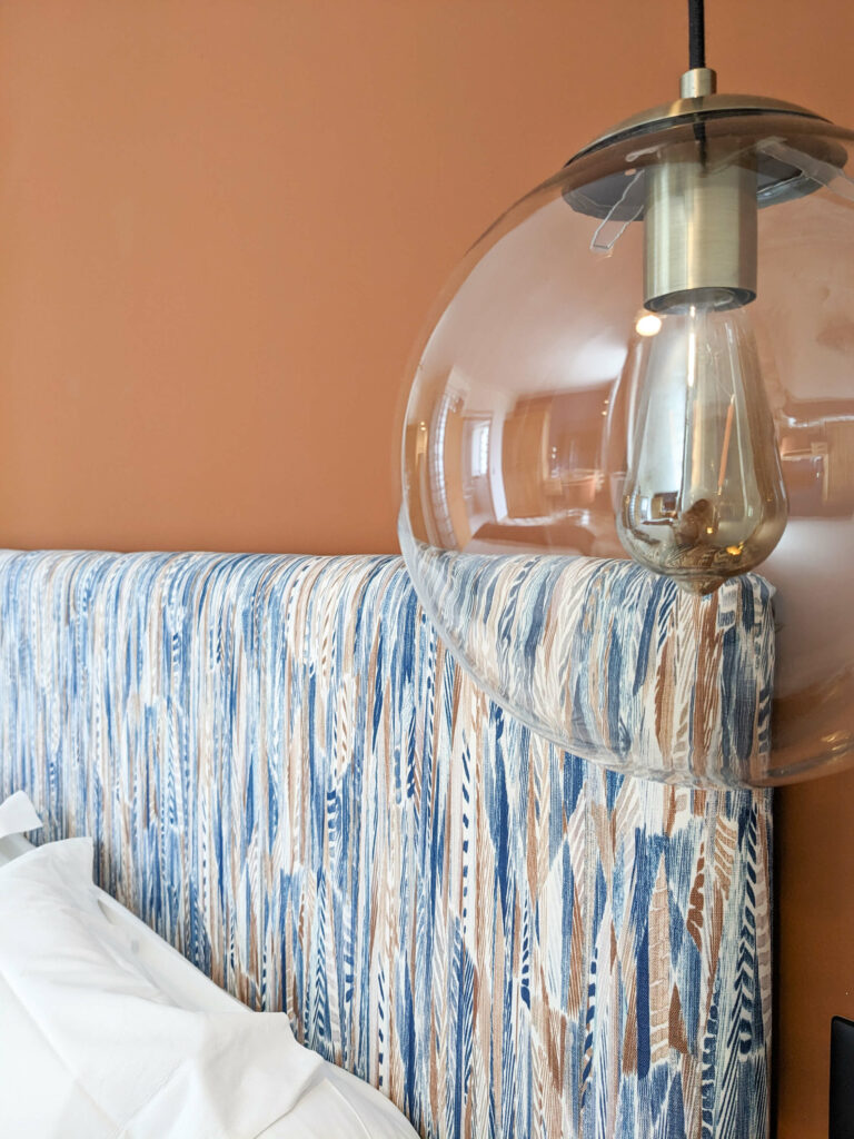 Rénovation d’une maison ancienne en chambre d’hôtes au Merlerault dans le perche normand, avec une tête de lit en tissu ethnique de plumes bleu et marron, un mur couleur terre cuite et des suspensions modernes en verre et laiton