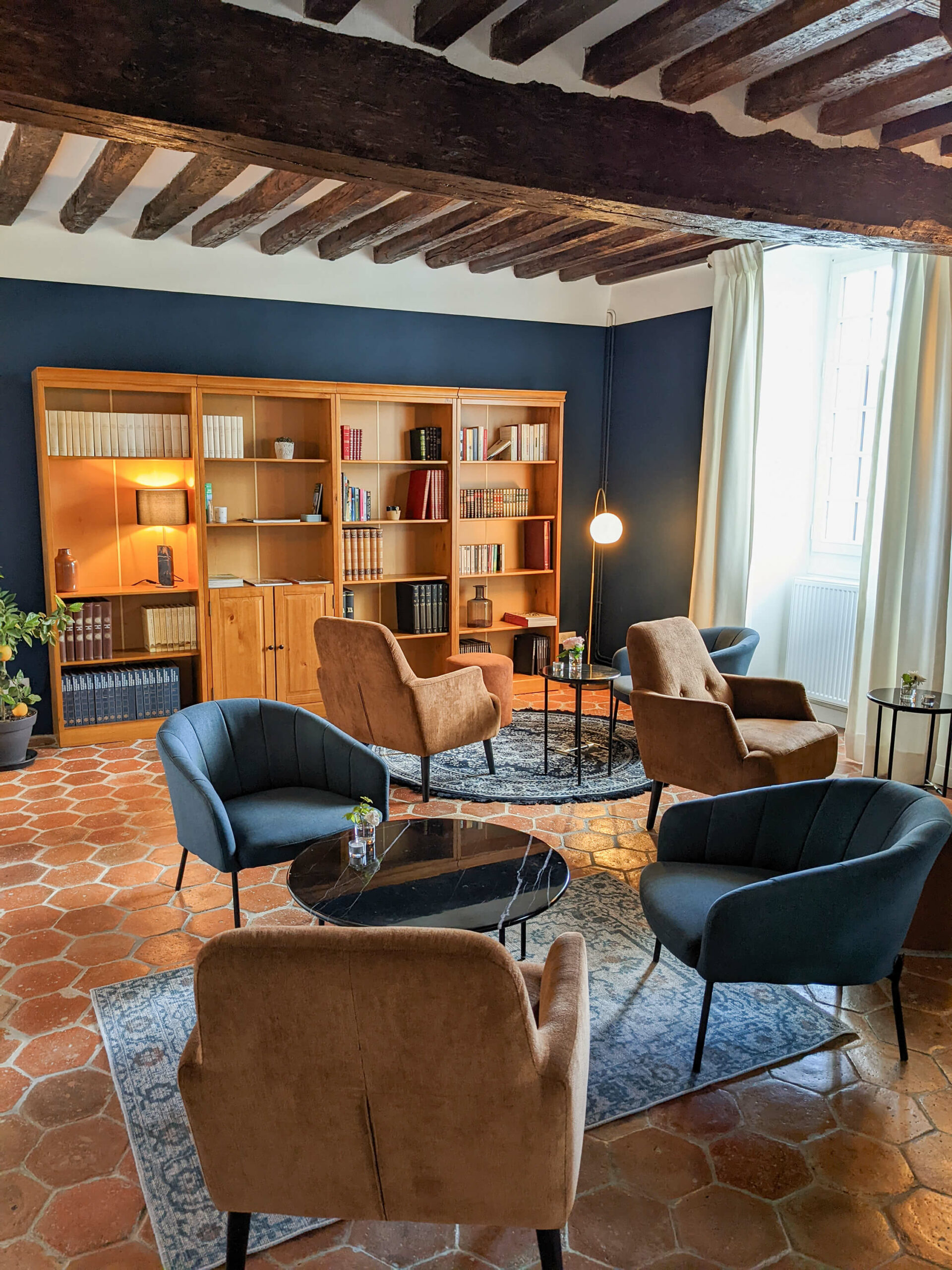Domaine de Prestal rénovation maisons d'hotes, boudoir avec salon bibliothèque Rénovation d’une maison ancienne en chambre d’hôtes au Merlerault dans le perche normand, espace salon avec poutres apparentes et tomettes au sol, des fauteuils dépareillés en velours terracotta et bleu, des tables en marbre noir, des tapis bleus et une grande bibliothèque en bois sur un mur bleu foncé