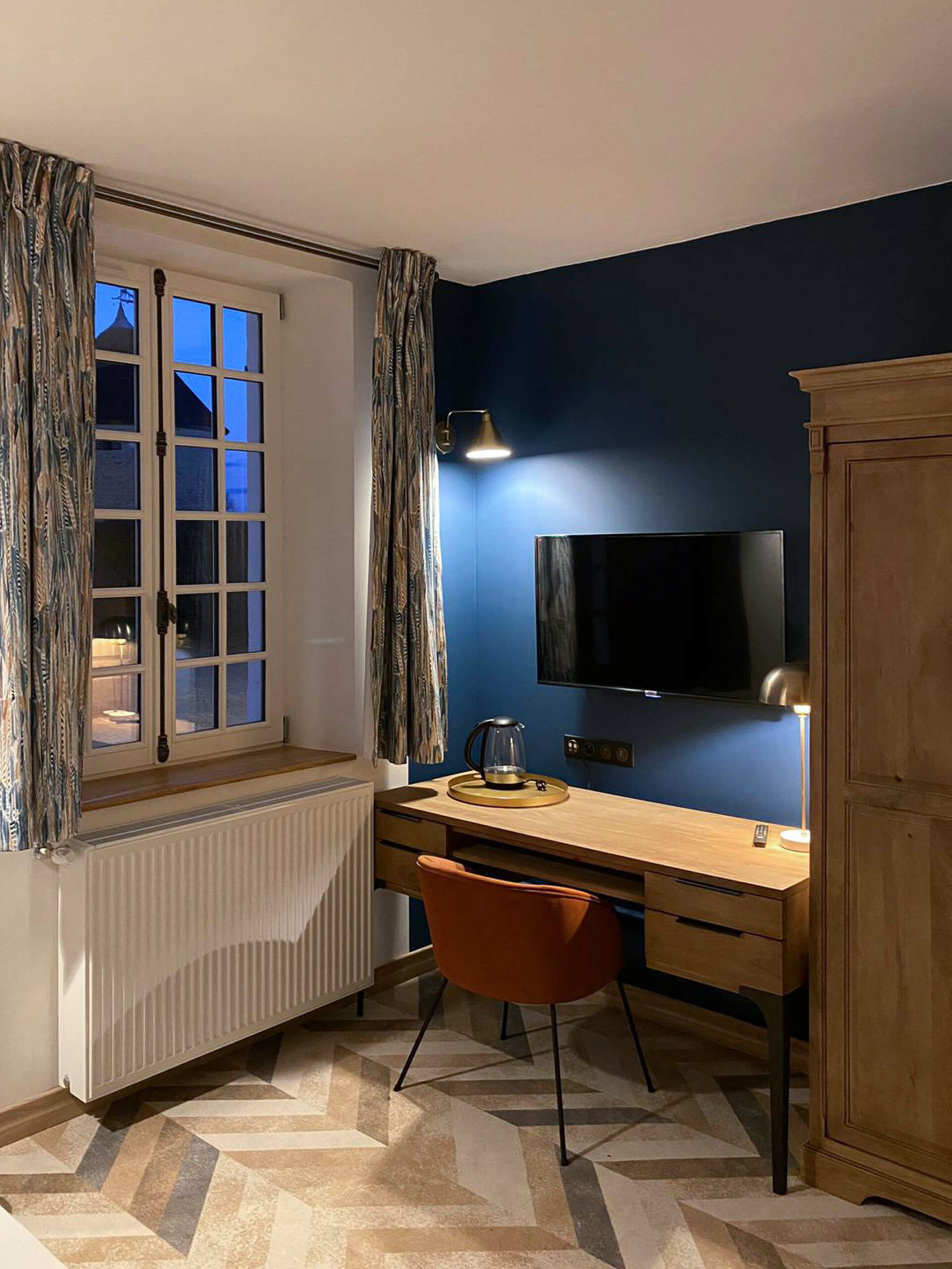 Domaine de Prestal rénovation maisons d'hotes chambre coin bureau avec mur bleu foncé, mobilier en bois massif et fauteuil terracotta Rénovation d’une maison ancienne en chambre d’hôtes au Merlerault dans le perche normand, esprit voyage, avec des rideaux en tissu ethnique de plumes bleu et marron, un mur bleu foncé, du mobilier en bois massif style colonial
