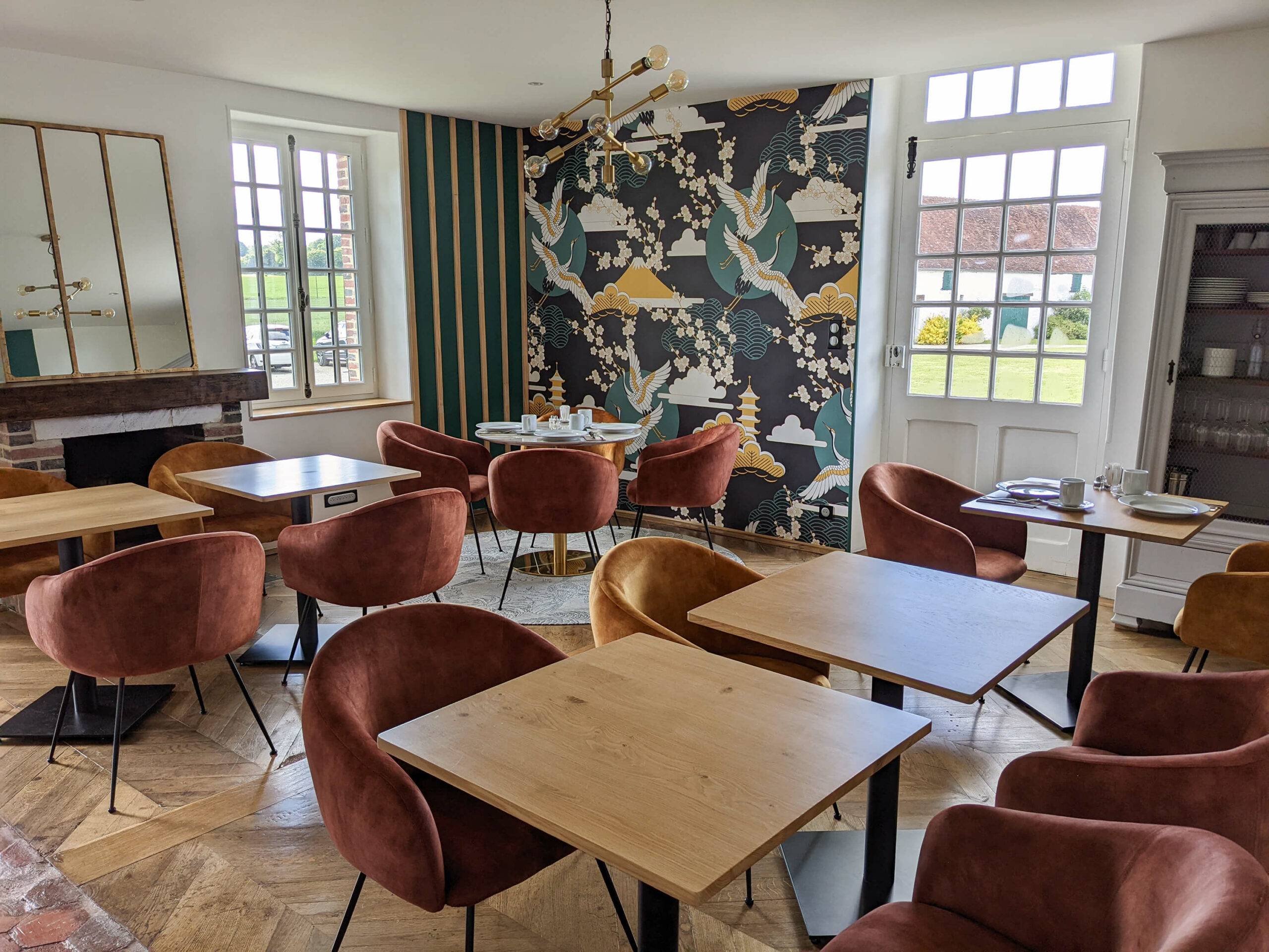 Domaine de Prestal rénovation maisons d'hotes, salle du petit déjeuner lumineuse et colorée avec un papier peint bucolique Rénovation d’une maison ancienne en chambre d’hôtes au Merlerault dans le perche normand, salle du petit déjeuner avec un papier peint japonisant, des tasseaux bois muraux, du parquet massif et des tomettes anciennes, tables en bois et fauteuils de table colorés, détails en laiton, cheminée en brique avec miroir laiton posé