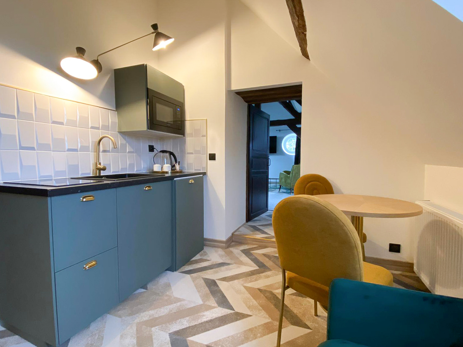 Rénovation d’une maison ancienne en chambre d’hôtes, kitchenette verte de gris avec poignées laiton et crédence en relief blanche Rénovation d’une maison ancienne en chambre d’hôtes au Merlerault dans le perche normand, esprit art déco, espace salon avec un canapé en velours vert, des chaises en velours doré, et kitchenette verte de gris avec poignées laiton et crédence en relief blanche