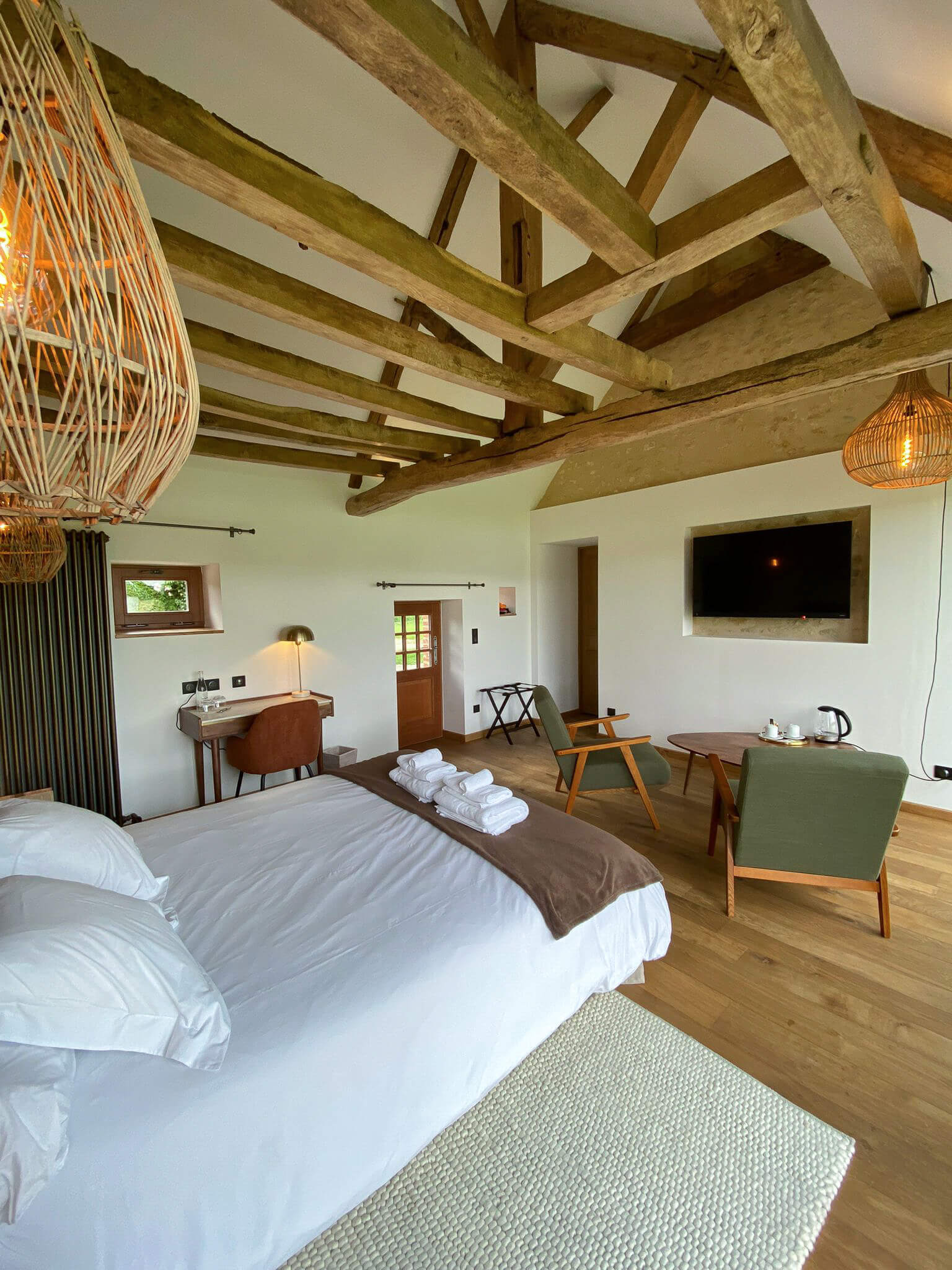 Réhabilitation d'un ancien four à pain en chambre d'hôtes chaleureuse avec poutres apparentes, mur en pierre et lumière naturelle, fauteuil en bois massif et tissu vert kaki dans le coin salon, au Merlerault dans le perche normand