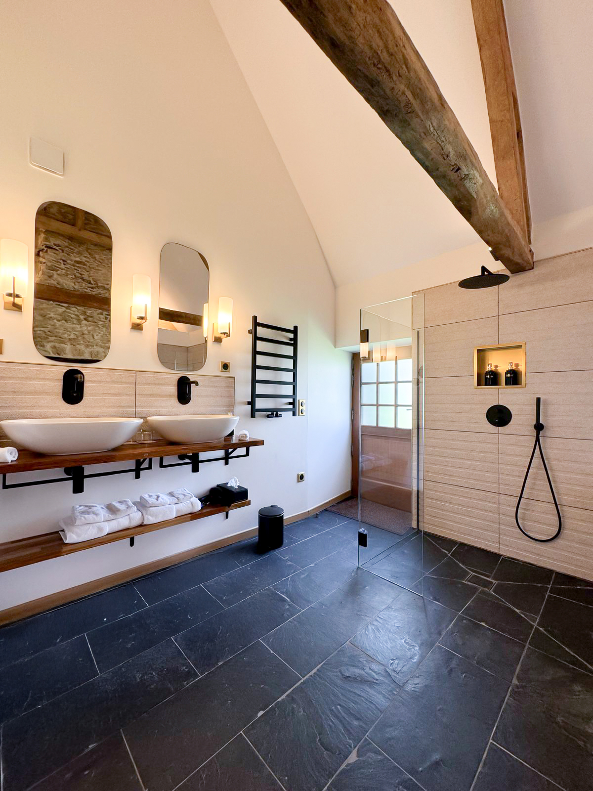 Réhabilitation d'un ancien four à pain en chambre d'hôtes chaleureuse avec poutres apparentes, mur en pierre et lumière naturelle, salle de bain balnéo avec grande douche et spa, plan en bois massif avec 2 vasques, sol ardoise, miroirs et luminaires laiton, au Merlerault dans le perche normand