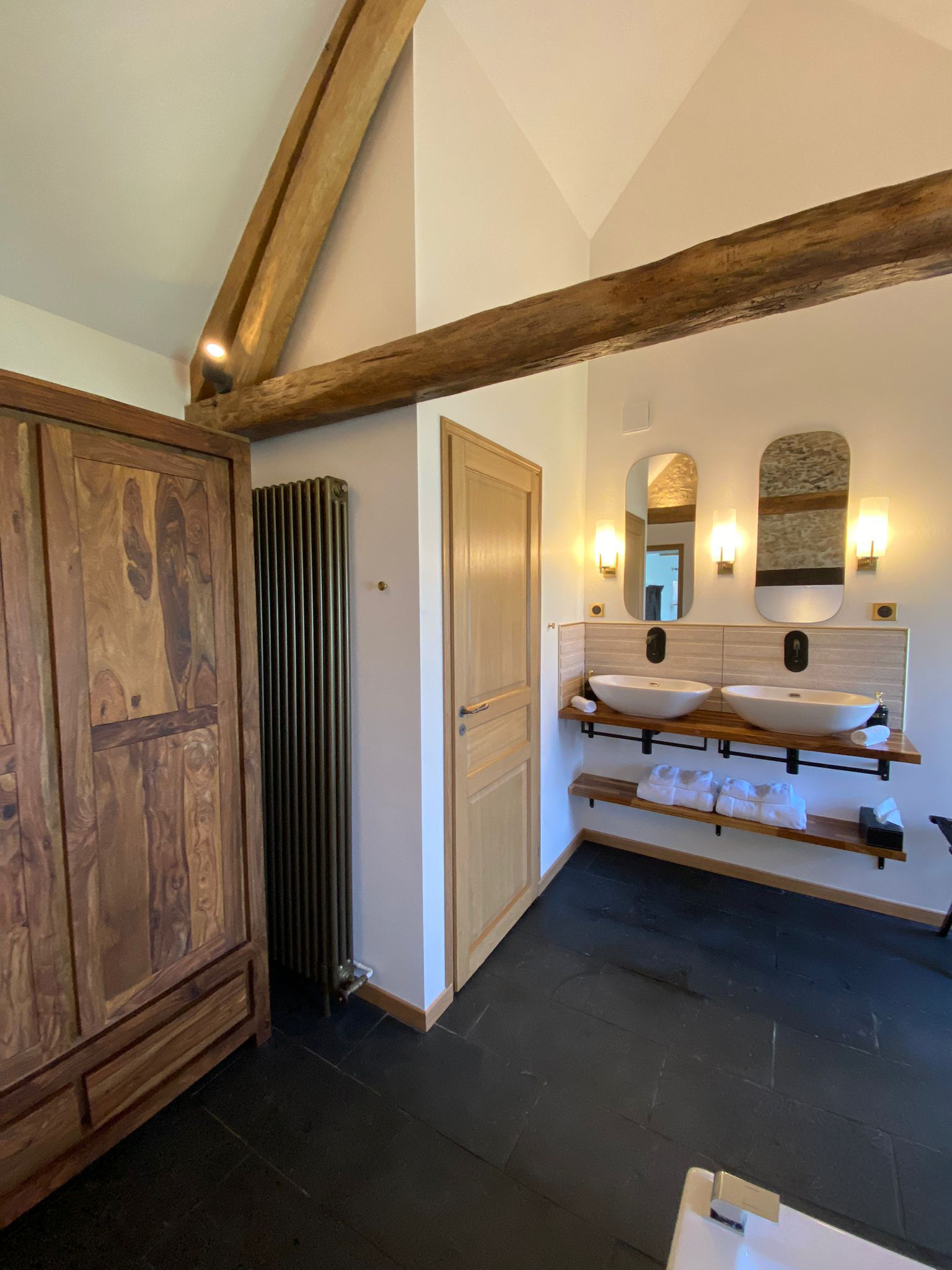 Réhabilitation d'un ancien four à pain en chambre d'hôtes chaleureuse avec poutres apparentes, mur en pierre et lumière naturelle, salle de bain balnéo avec grande douche et spa, plan en bois massif avec 2 vasques, sol ardoise, miroirs et luminaires laiton, au Merlerault dans le perche normand