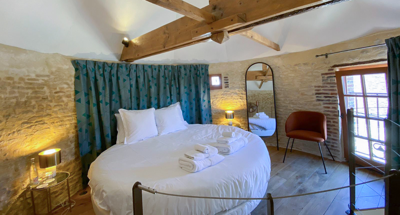 Domaine de Prestal rénovation ancien pigeonnier, chambre avec lit rond, charpente apparente et mur de pierre Réhabilitation d'un ancien pigeonnier en chambre d'hôtes, avec poutres apparentes et murs en pierres, lit rond, tête de lit en rideaux à motifs géométriques, détails cuivrés, au Merlerault dans le perche normand