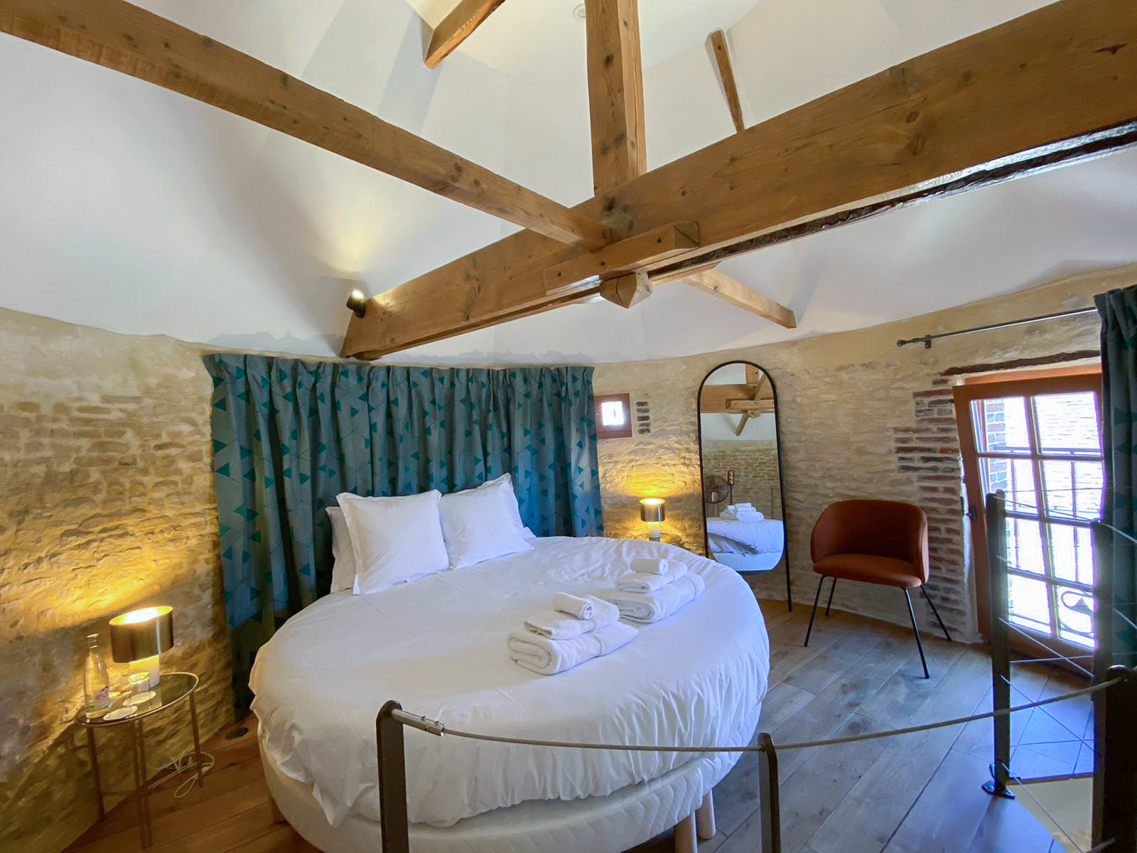 Domaine de Prestal rénovation ancien pigeonnier, chambre avec lit rond, charpente et mur de pierre Réhabilitation d'un ancien pigeonnier en chambre d'hôtes, avec poutres apparentes et murs en pierres, lit rond, tête de lit en rideaux à motifs géométriques, détails cuivrés, au Merlerault dans le perche normand