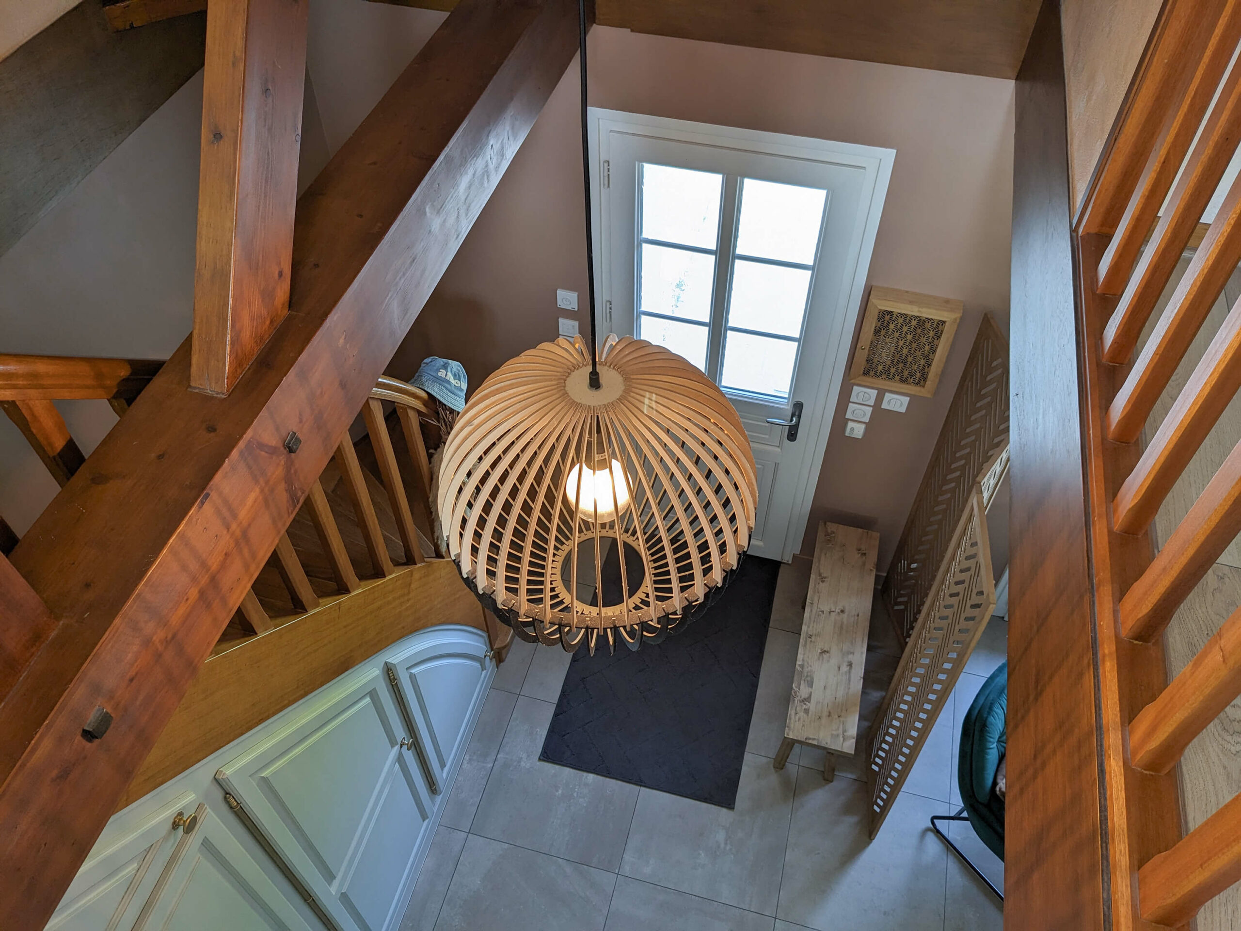 Rénovation d’une maison familiale en campagne essonnienne à Chalo-Saint-Mars, vue sur l’entrée avec poutres en bois apparentes, suspension boule en bois, paravent et banc en bois, mur terracotta et fauteuil en velours vert sapin