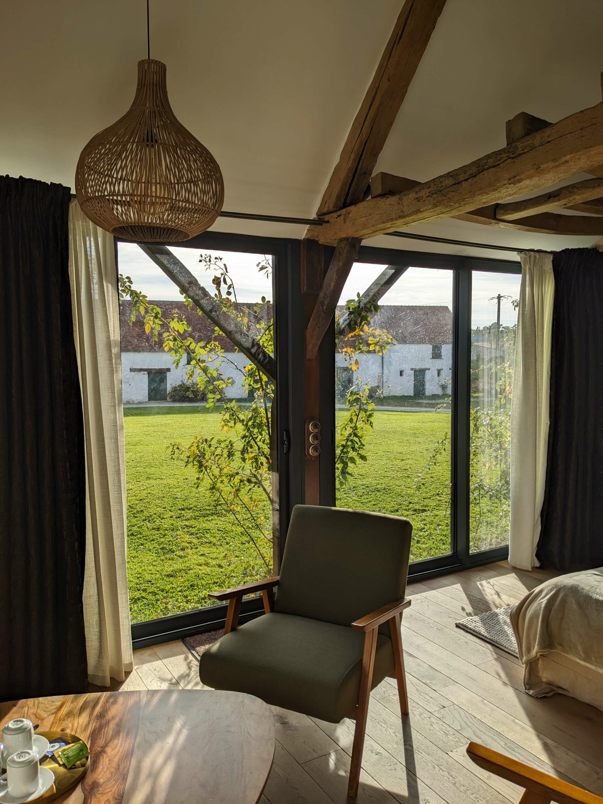 Réhabilitation d'un ancien four à pain en chambre d'hôtes chaleureuse avec poutres apparentes, mur en pierre et lumière naturelle, fauteuil en bois massif et tissu vert kaki dans le coin salon, au Merlerault dans le perche normand