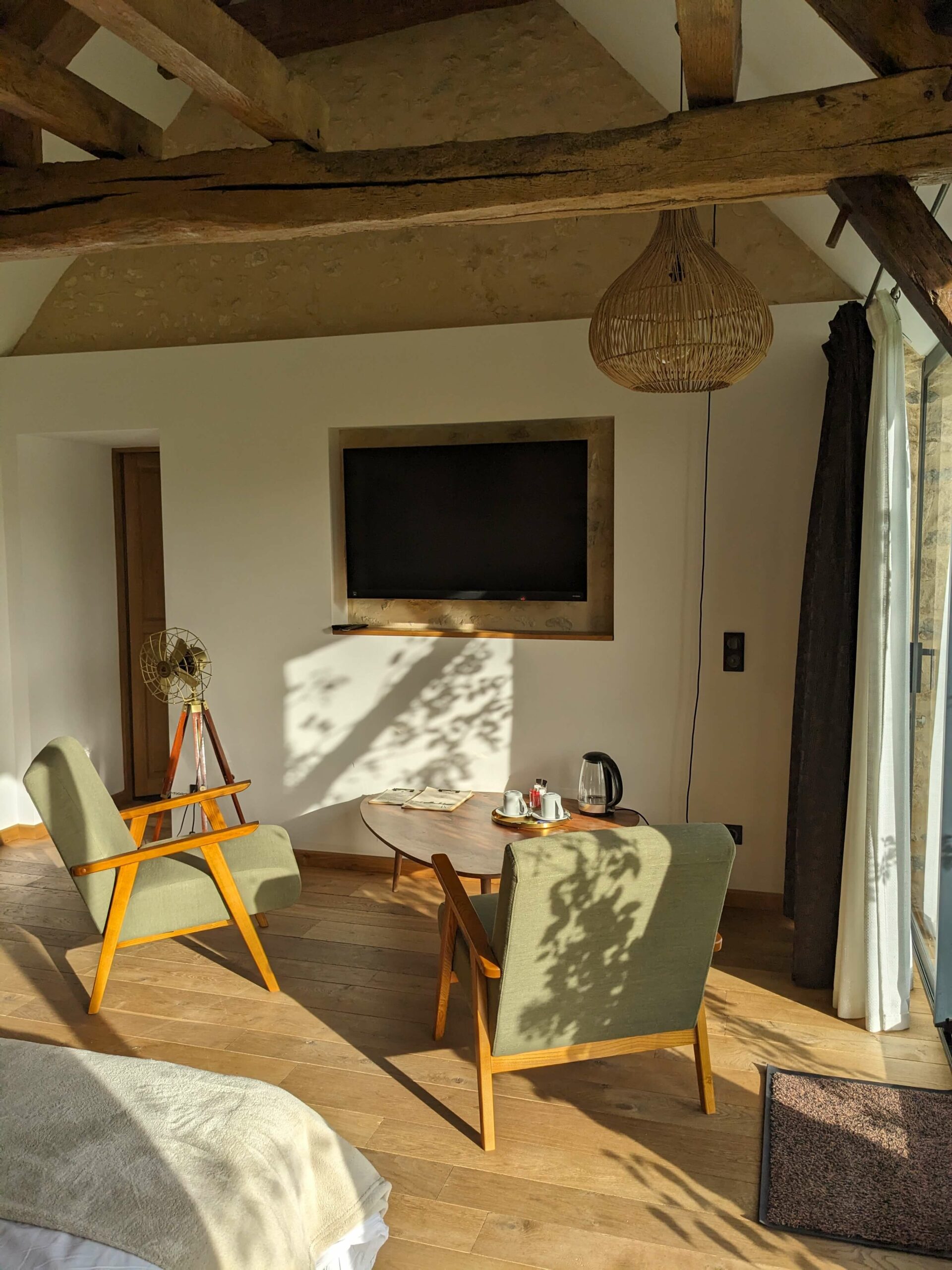 Réhabilitation d'un ancien four à pain en chambre d'hôtes chaleureuse avec poutres apparentes, mur en pierre et lumière naturelle, fauteuil en bois massif et tissu vert kaki dans le coin salon, au Merlerault dans le perche normand