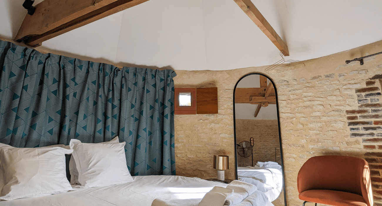 Réhabilitation d'un ancien pigeonnier en chambre d'hôtes, avec poutres apparentes et murs en pierres, lit rond, tête de lit en rideaux à motifs géométriques, détails cuivrés, au Merlerault dans le perche normand
