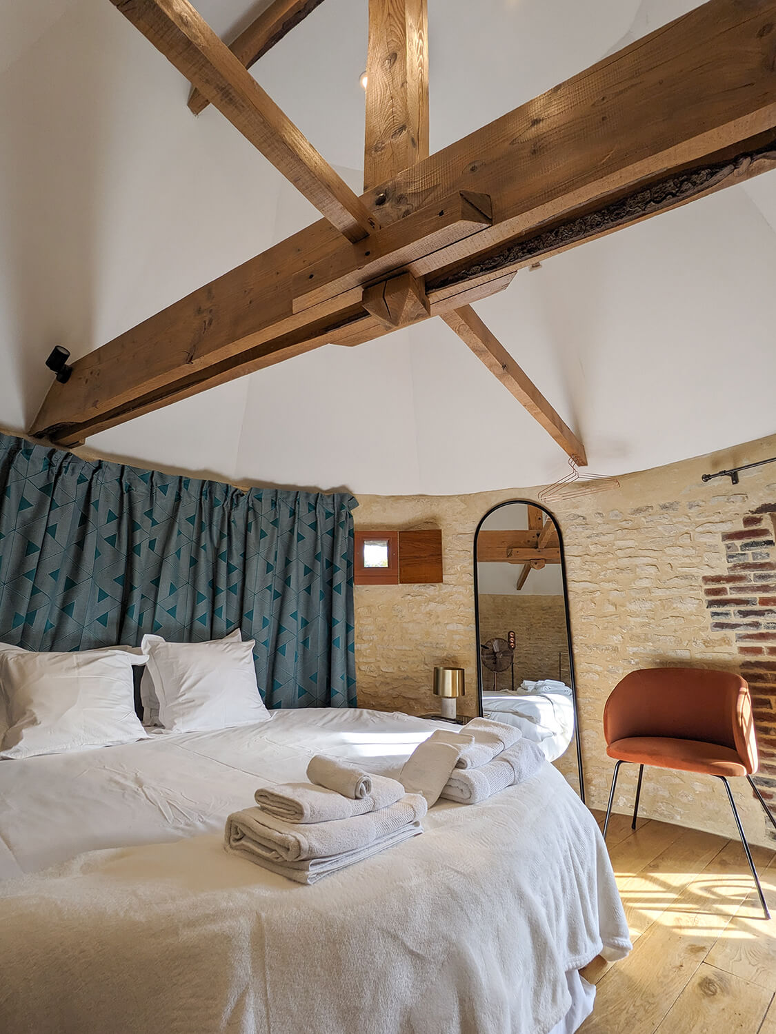 Réhabilitation d'un ancien pigeonnier en chambre d'hôtes, avec poutres apparentes et murs en pierres, lit rond, tête de lit en rideaux à motifs géométriques, détails cuivrés, au Merlerault dans le perche normand